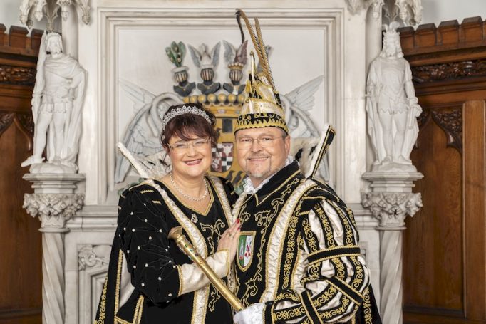 Paar in festlicher, traditioneller Kleidung vor einem Wappen und Säulen.